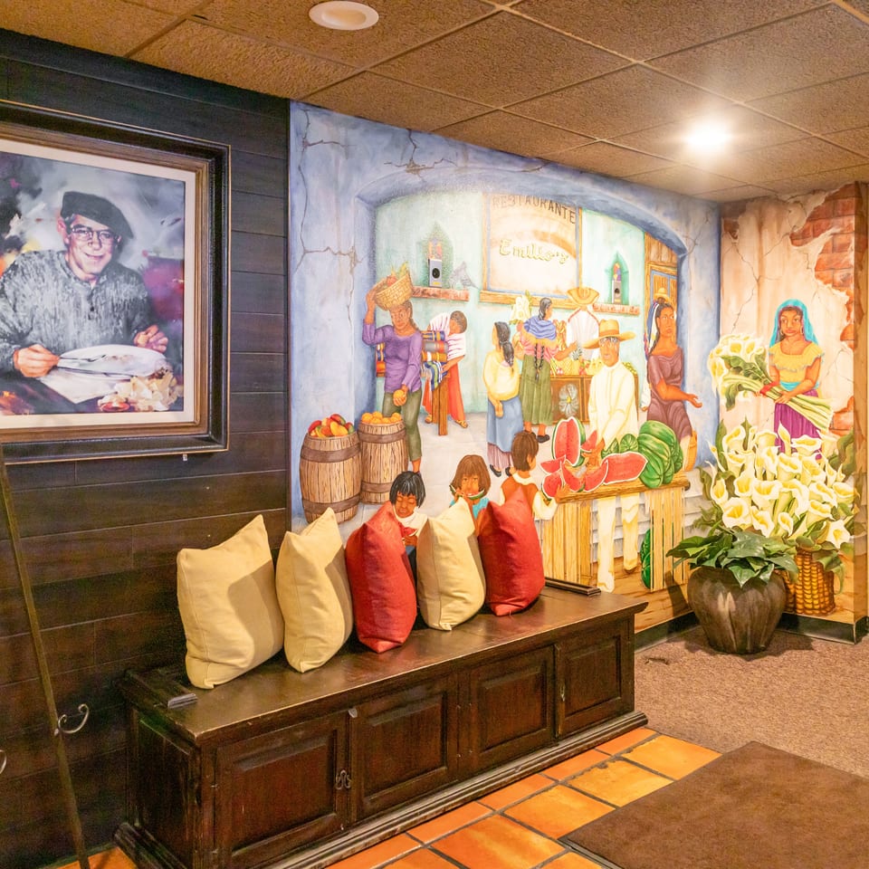 Restaurant interior featuring a colorful mural of a market scene, floral display, portrait, and cushioned wooden bench.