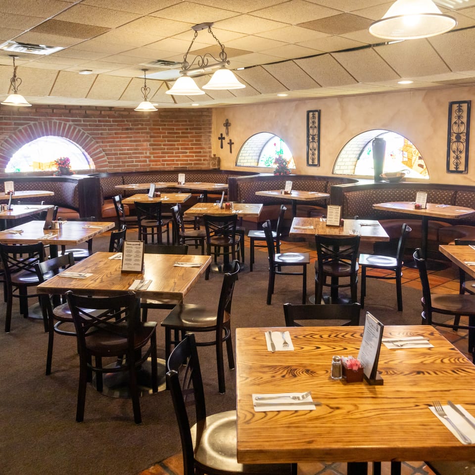 Spacious restaurant dining room with wood tables, dark chairs, cushioned booths, brick walls, arched windows, and pendant lighting.