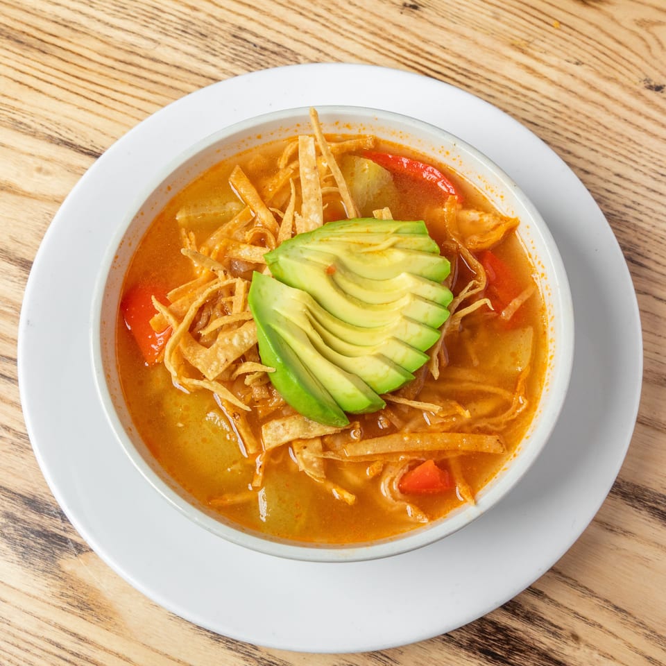 Bowl of orange soup topped with sliced avocado, crispy tortilla strips, tomatoes, and vegetables on a white plate.