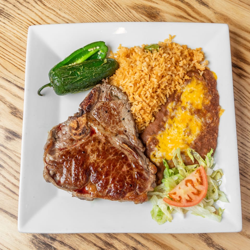 Grilled steak served with Mexican rice, cheese-covered beans, roasted jalapeños, and a lettuce-tomato salad on a white plate.