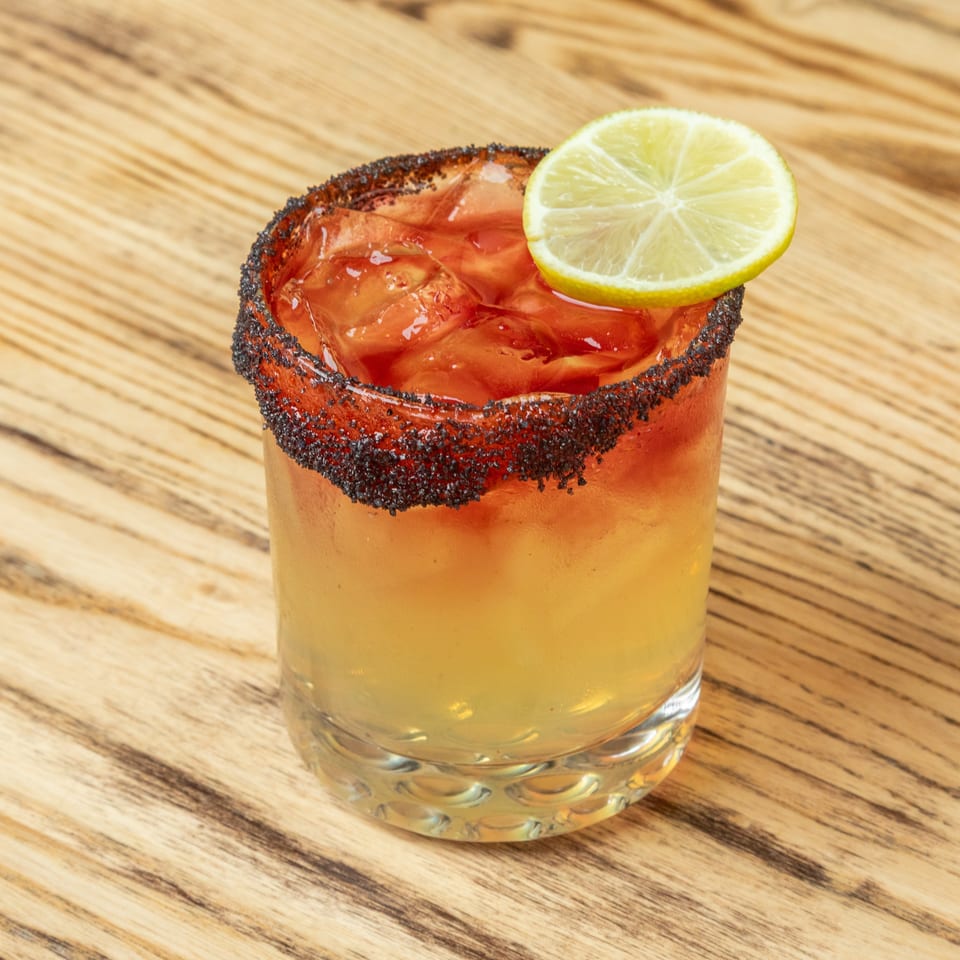 Golden cocktail with ice, a chili-salt rim, and lime garnish served in a glass on a wooden table.