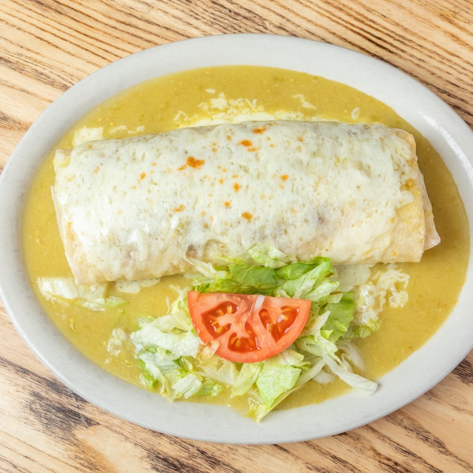 Cheese-covered burrito served in green sauce with shredded lettuce and tomato on a white plate.