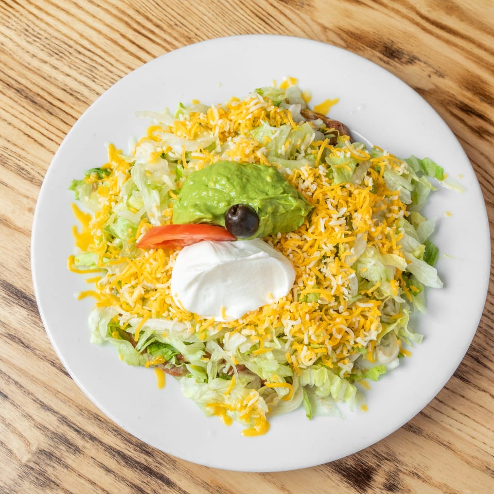 Taco salad topped with shredded lettuce, cheddar cheese, guacamole, sour cream, tomato, and black olive on a white plate.