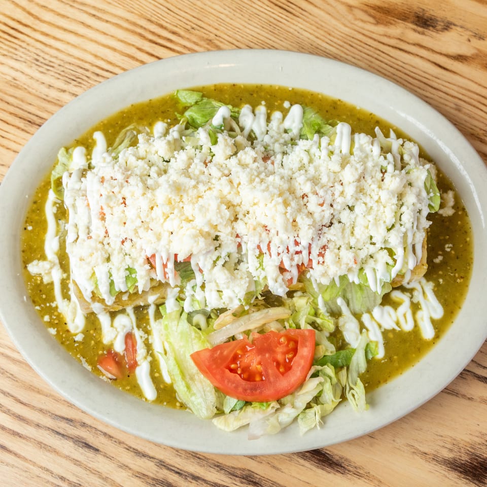 Mexican dish topped with shredded lettuce, tomatoes, crumbled cheese, and crema over green salsa on an oval plate.