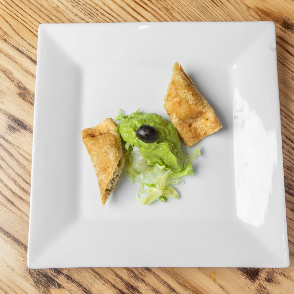 Crispy triangular pastries served with lettuce, guacamole, and a black olive on a white plate.