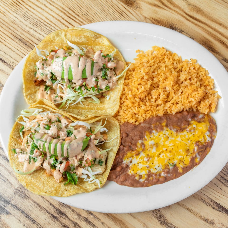 Mexican plate with two saucy tacos, orange rice, and refried beans topped with melted cheese on a wooden table.