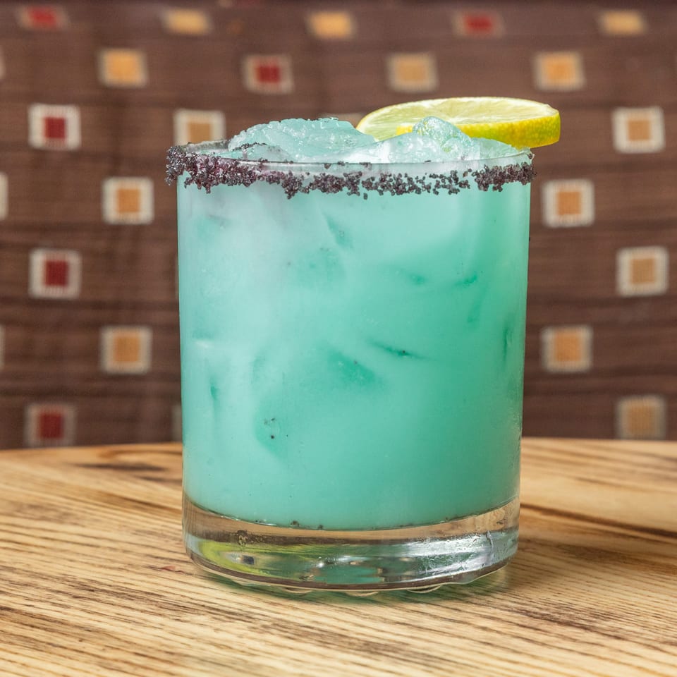 Bright turquoise cocktail with crushed ice, black salt rim, and lime wedge served in a glass on a wooden table.