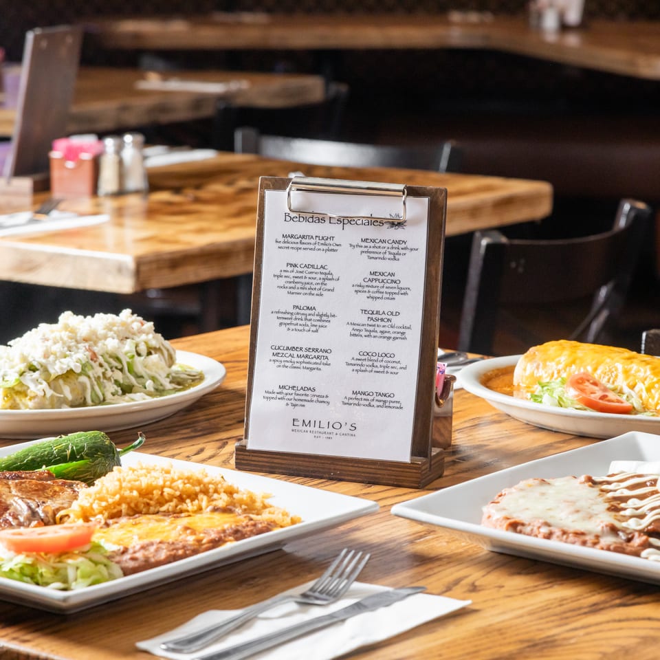 Mexican restaurant table displaying assorted plated dishes and a menu of special beverages in a warmly lit dining room.