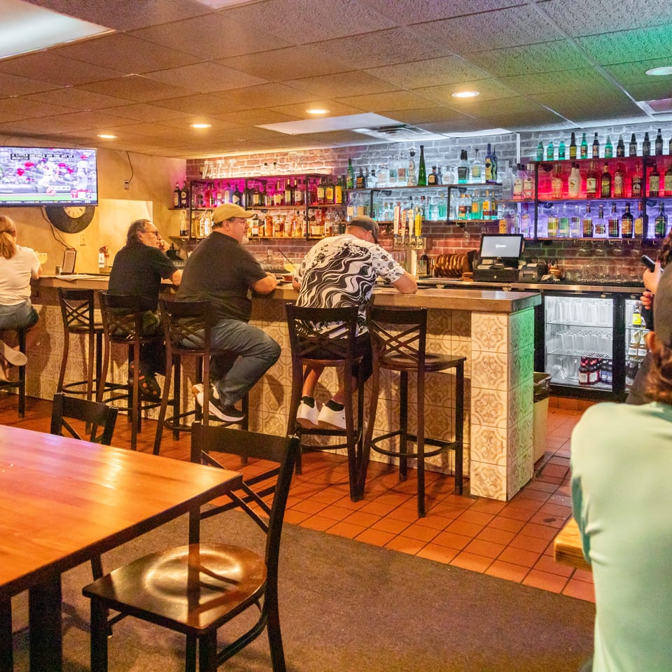 Busy restaurant bar with patrons seated on stools, illuminated liquor shelves, tiled counter, and televisions overhead.
