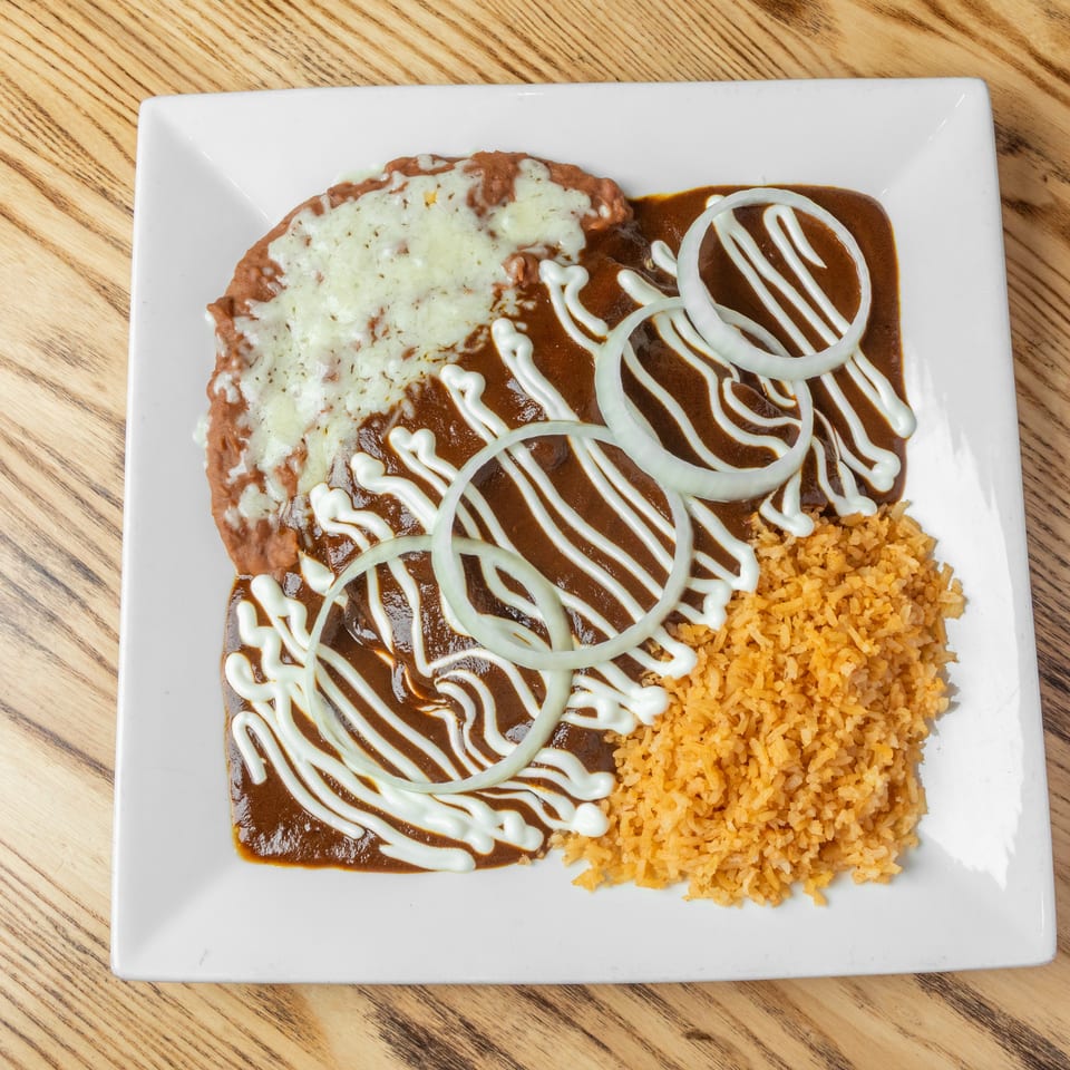 Mexican-style plate with cheese-covered refried beans, enchiladas topped with brown sauce, crema, and onions, served with Spanish rice.