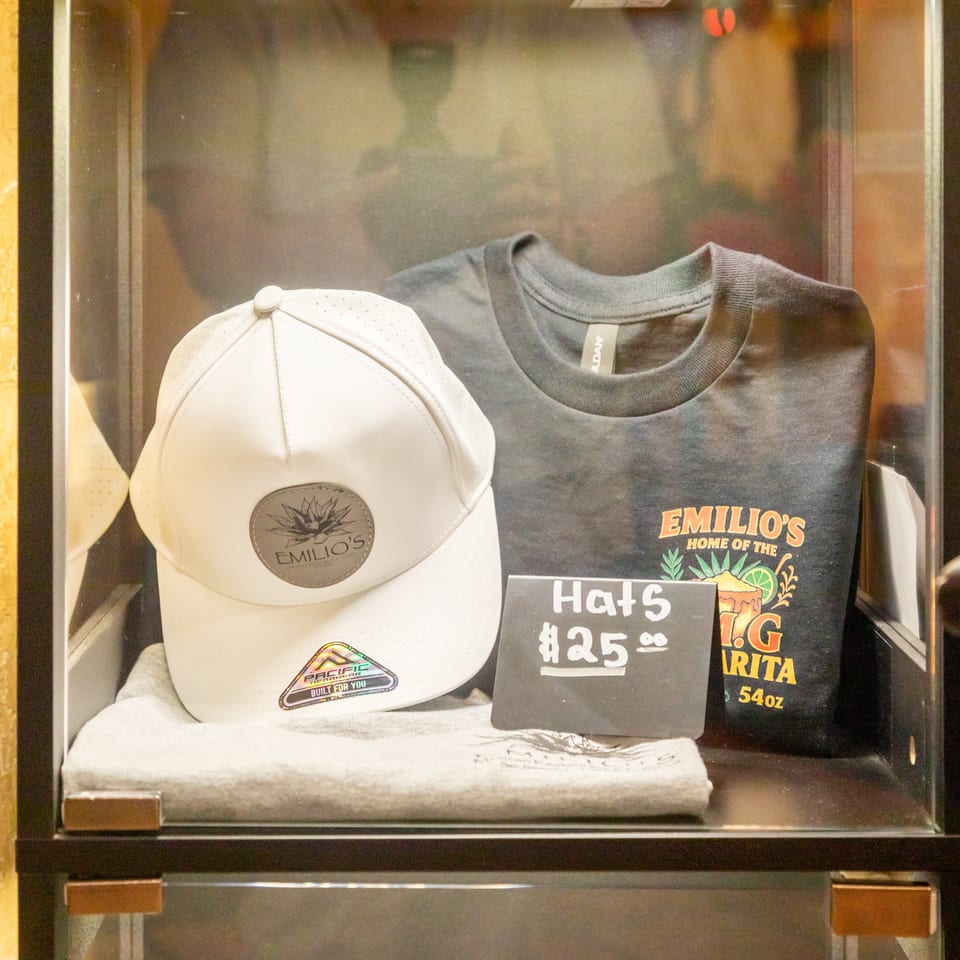 Emilio’s merchandise display featuring a white cap, gray T-shirt, folded shirt, and chalkboard advertising hats for $25.
