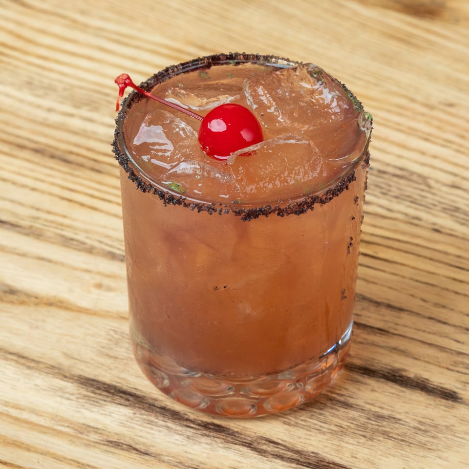Amber cocktail with ice, a cherry garnish, and dark sugar rim served in a tumbler on a wooden table.