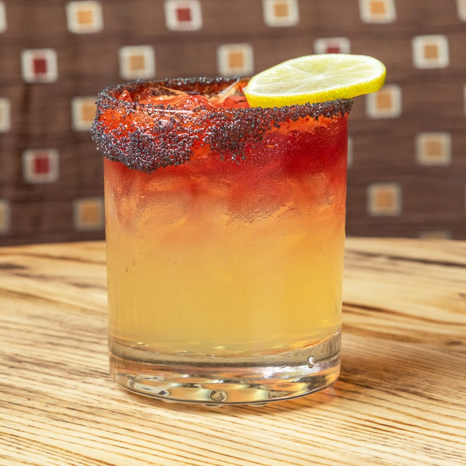 Layered orange-red cocktail in a glass with a black-salt rim and lime garnish, resting on a wooden bar.