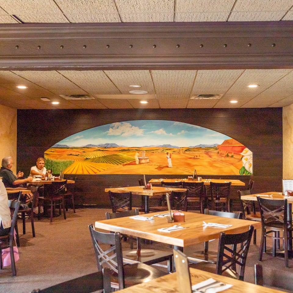 Warmly lit restaurant dining room with wooden tables, seated diners, and a colorful mural depicting farmland and mountains.