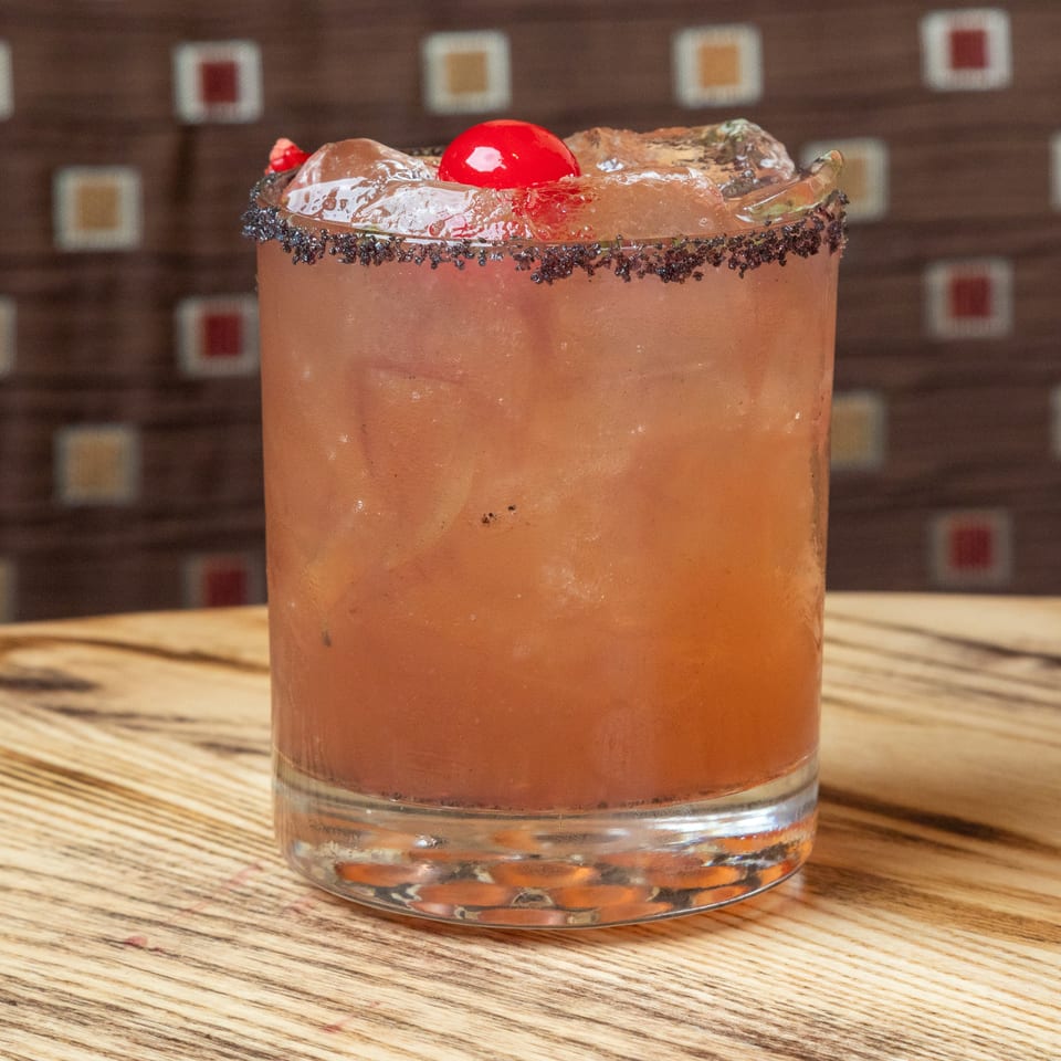 Orange cocktail in a rocks glass with a black salt rim, ice, cherry, and lime garnish on a wooden bar.