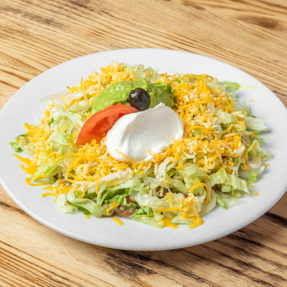Taco salad with shredded lettuce, cheese, tomato, avocado, black olive, rice, and sour cream on a white plate.