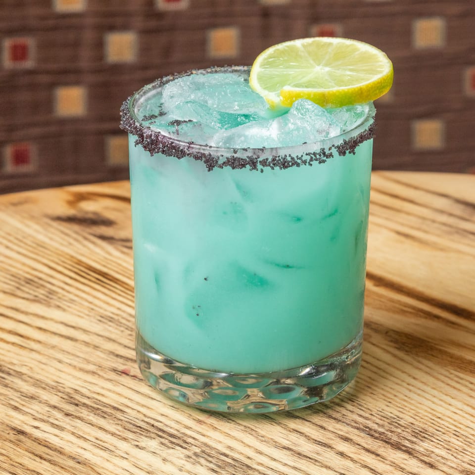 Blue cocktail with crushed ice, a dark salt rim, and lime garnish on a wooden restaurant table.